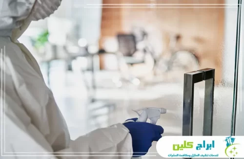 شركة تعقيم بجازان 0550234190 ابراج كلين يظهر أحد المختصين مرتدياً بدلة واقية كاملة مع قناع، وهو يقوم برش مواد معقمة على الأسطح الزجاجية والنوافذ