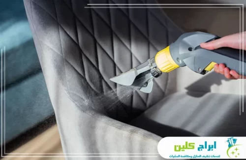 شركة غسيل مجالس بالدمام 0550234190 ابراج كلين رش سائل التنظيف بقوة على ظهر كرسي رمادي بتصميم عصري لتطهيره.